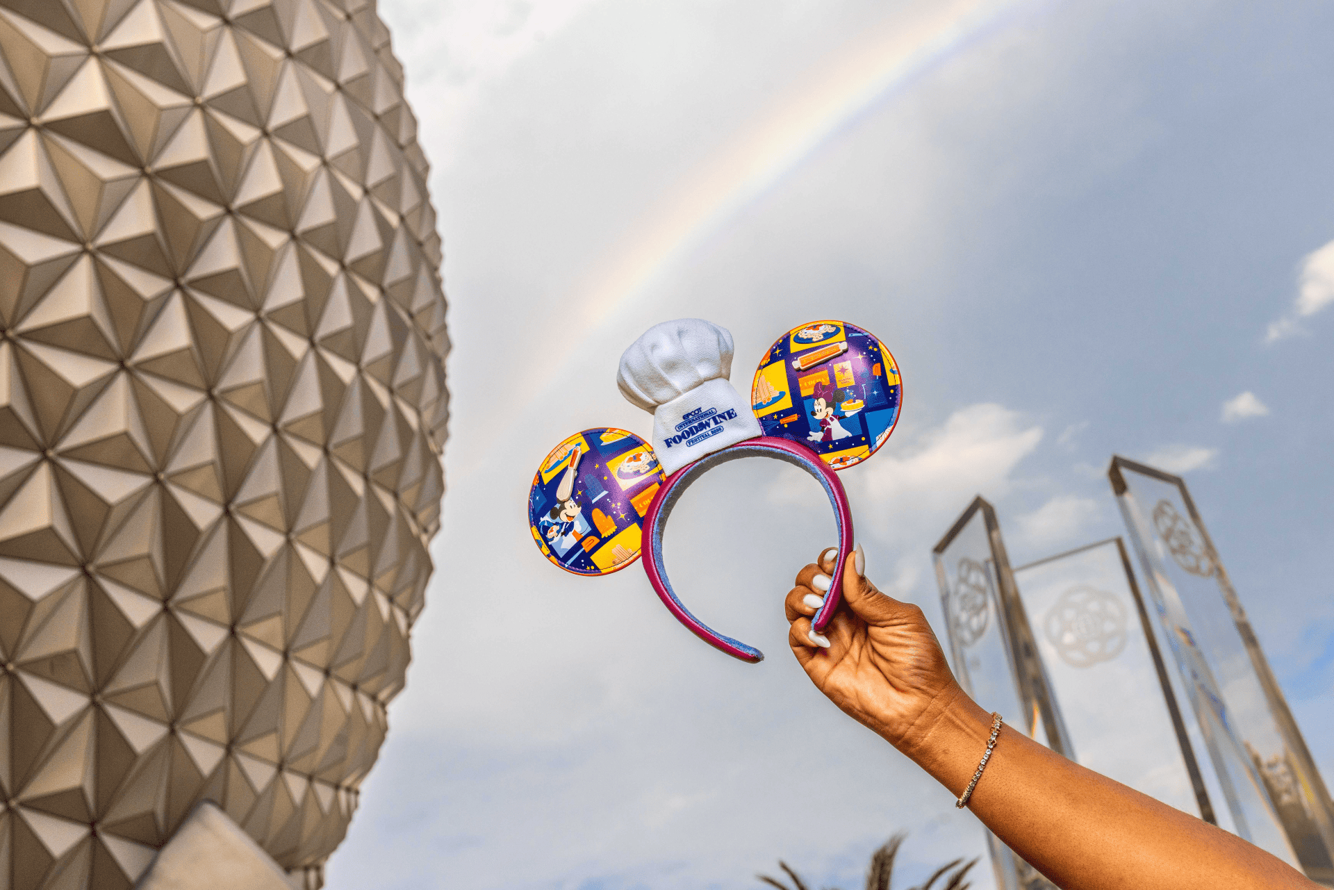 Person holding Mickey Mouse ears with Epcot sphere in background and rainbow.