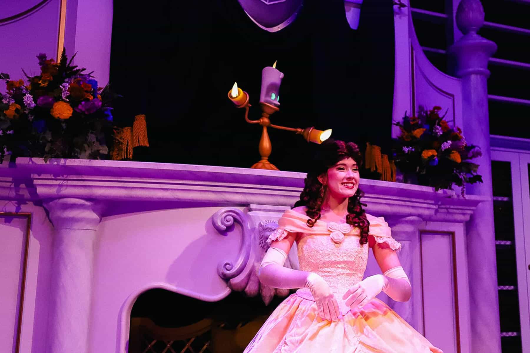 Belle smiles to guests with Lumiere on the mantle behind her. 