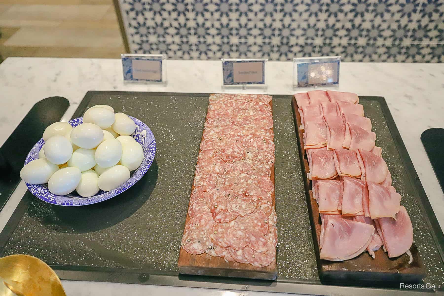 a bowl of hard-boiled eggs with platters of salami and ham 