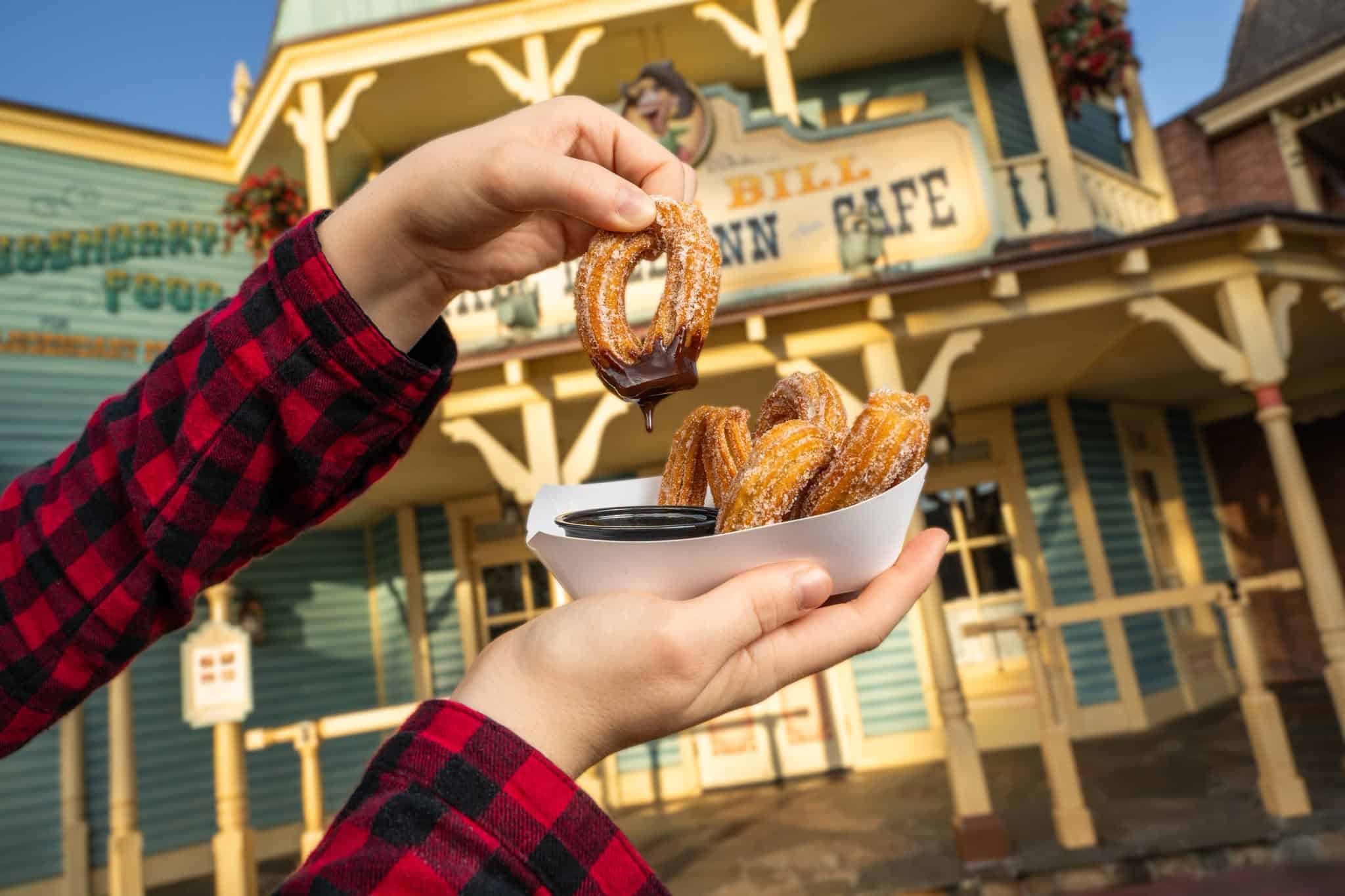 a hand holding an order of churros with chocolate dipping sauce in front of Pecos Bill