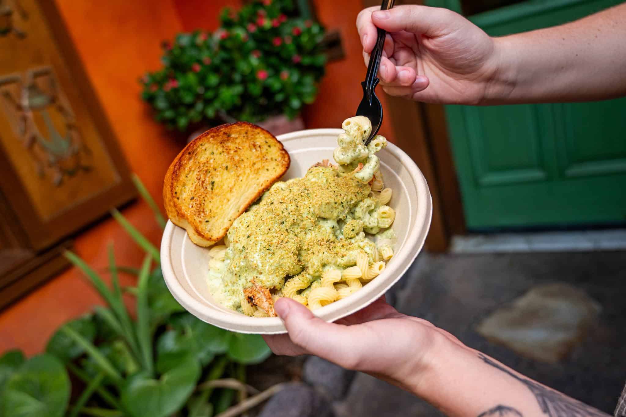 a hand holding a bowl of creamy pesto chicken pasta with toast