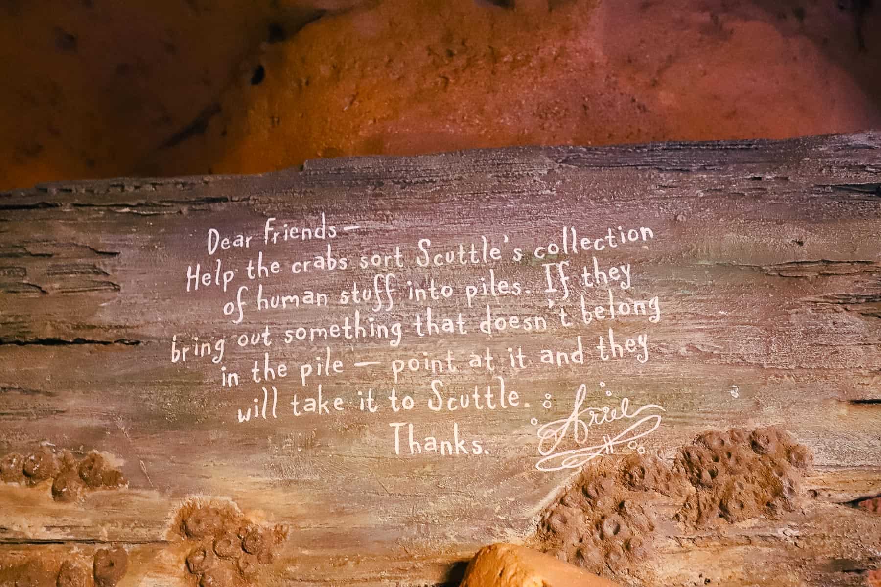 Interactive queue sign reads, "Dear Friends, Help the crabs sort Scuttle's collection of human stuff into piles. If they bring out something that doesn't belong in the pile, point at it and they will take it to Scuttle. Thanks, Ariel 