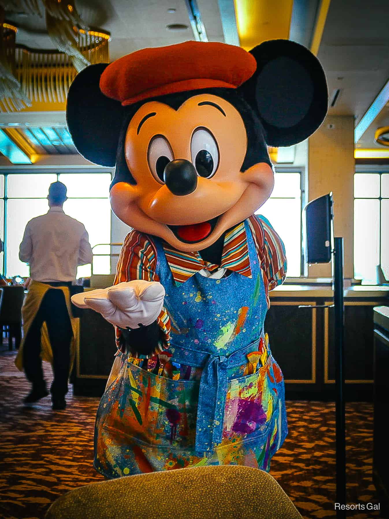 Mickey Mouse poses with his hand out in his painter's apron. 