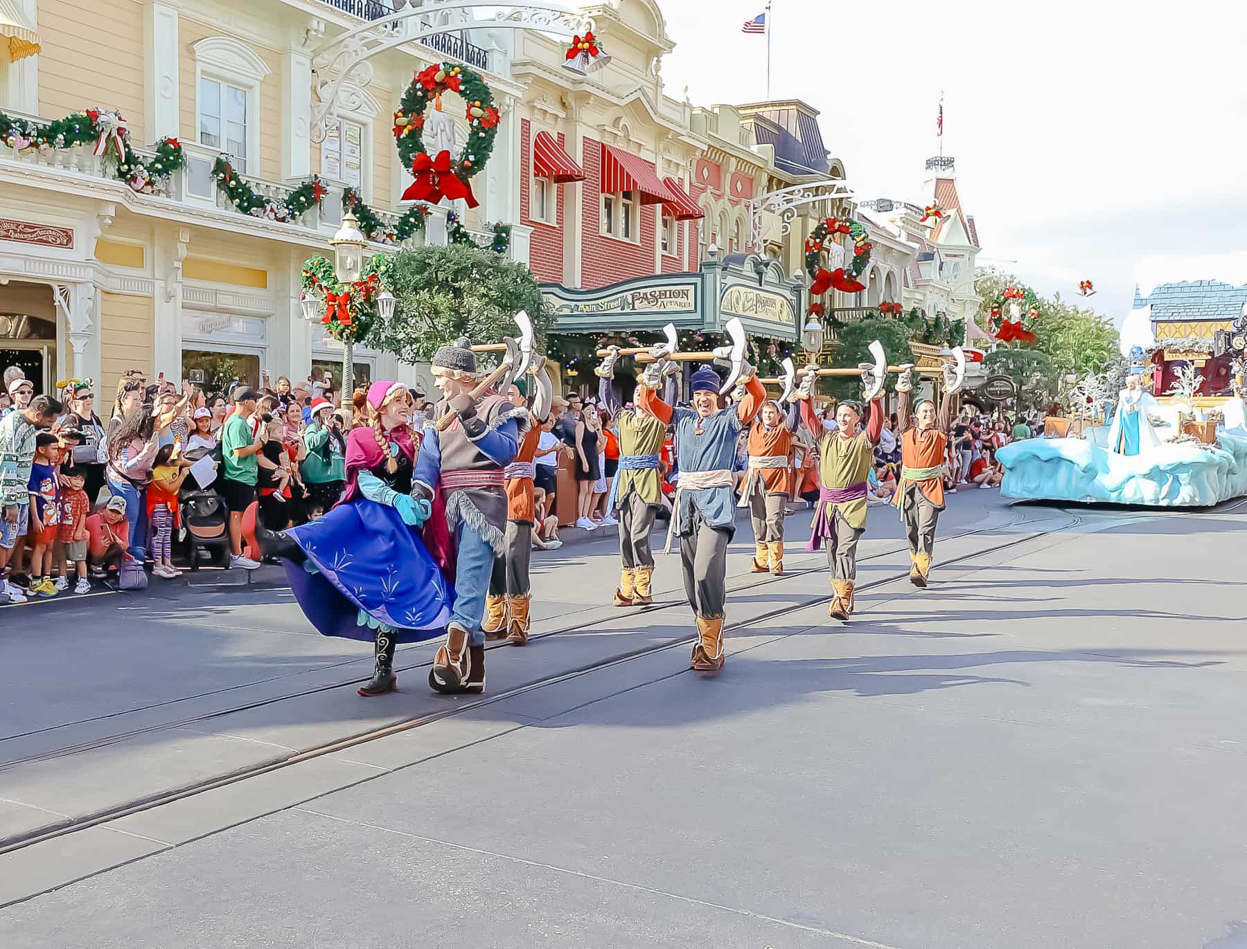 Anna and Kristoff walking in front of a group of icemen in the Once Upon a Christmastime parade. 