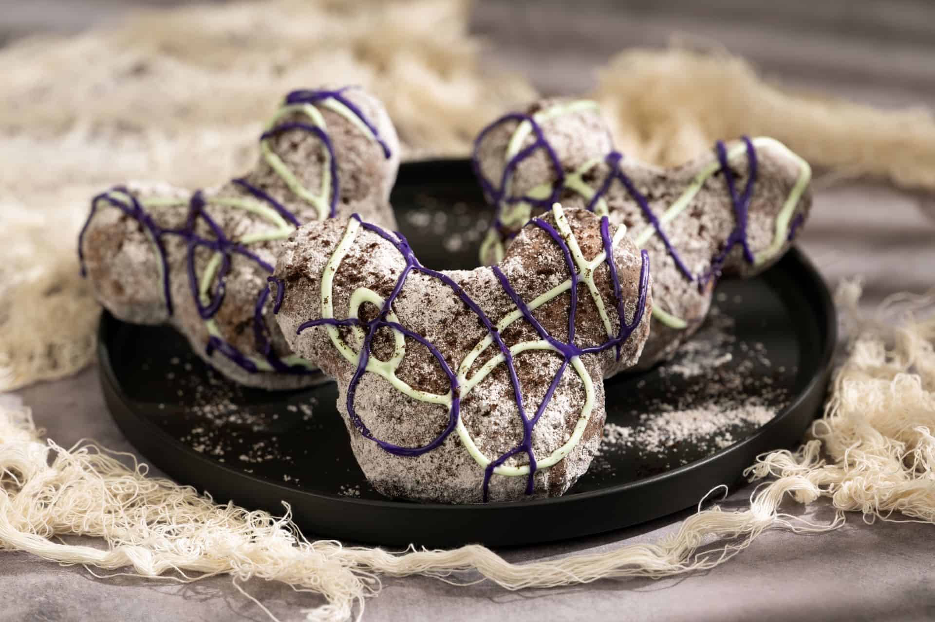 a platter of beignets with a grey color like cob webs topped with white and purple icing