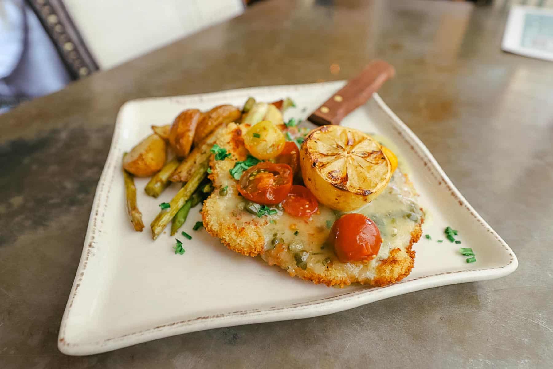 An order of Chicken Paillard topped with a roasted lemon with a side of asparagus and potatoes. 