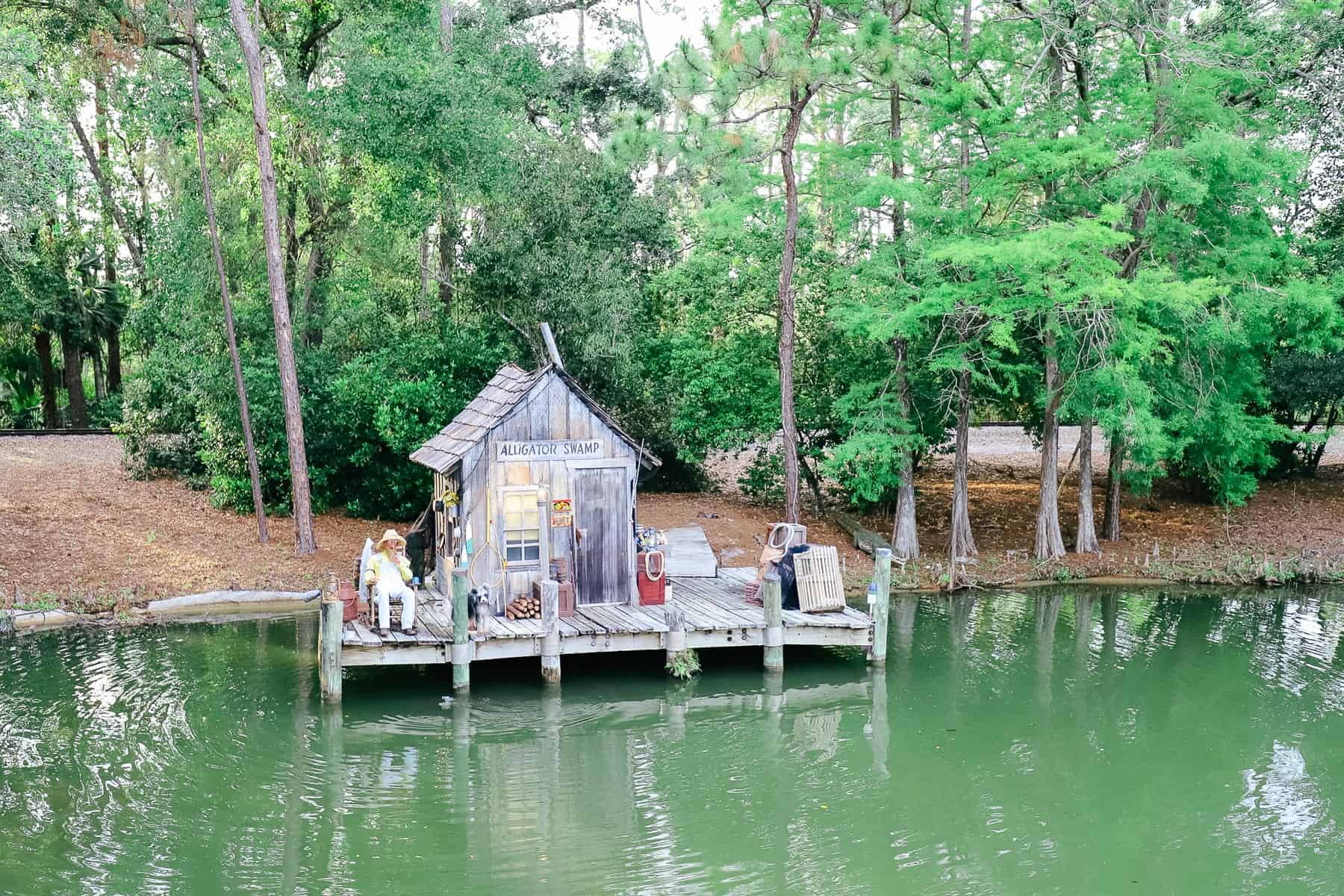 A scene along the river that shows a man fishing in front of a shack with a sign that says Alligator Swamp. 