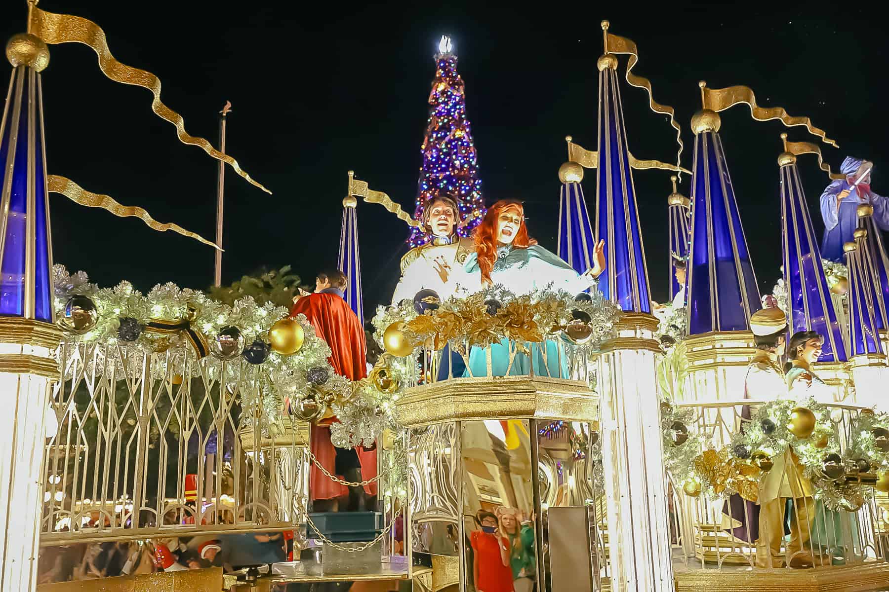 Prince Eric and Ariel wave with the Christmas tree behind them. 