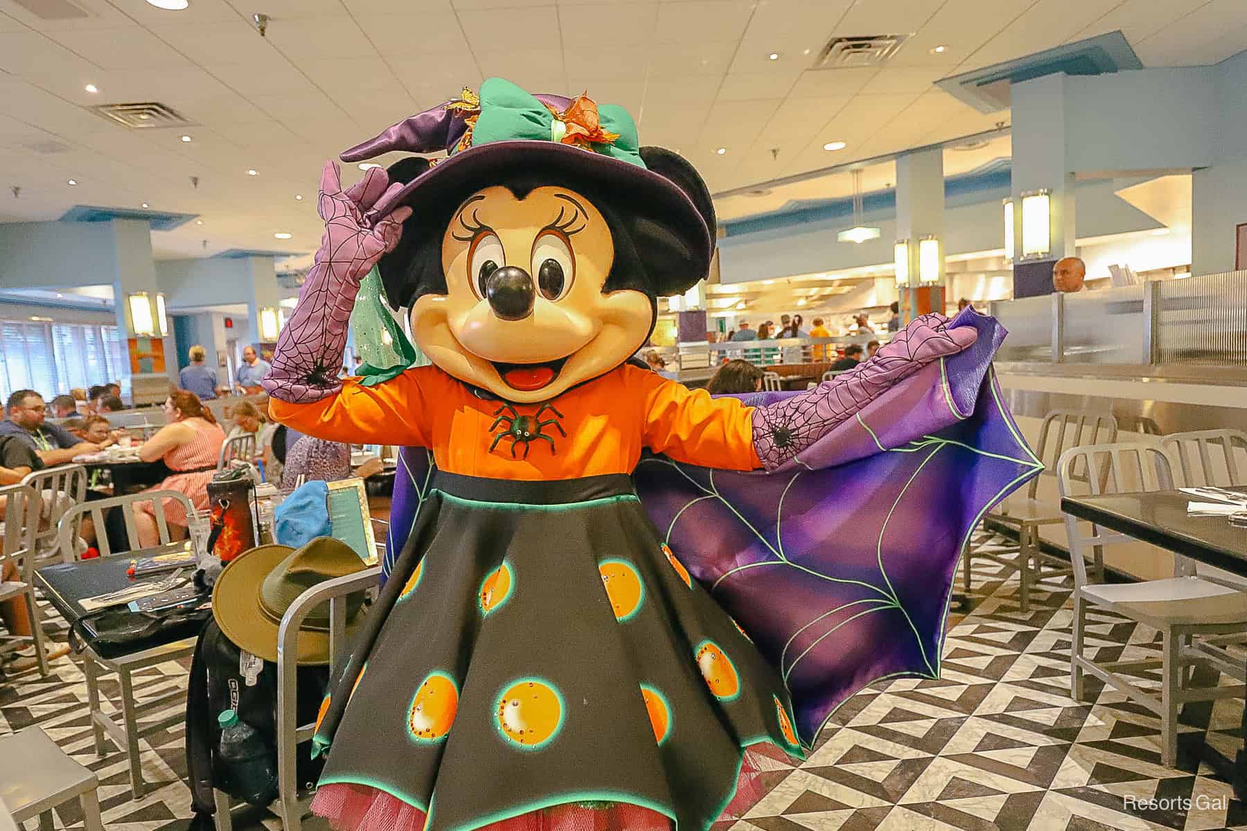 Minnie Mouse has a witches hat and is wearing an orange and purple outfit with a cape that matches Mickey's. 