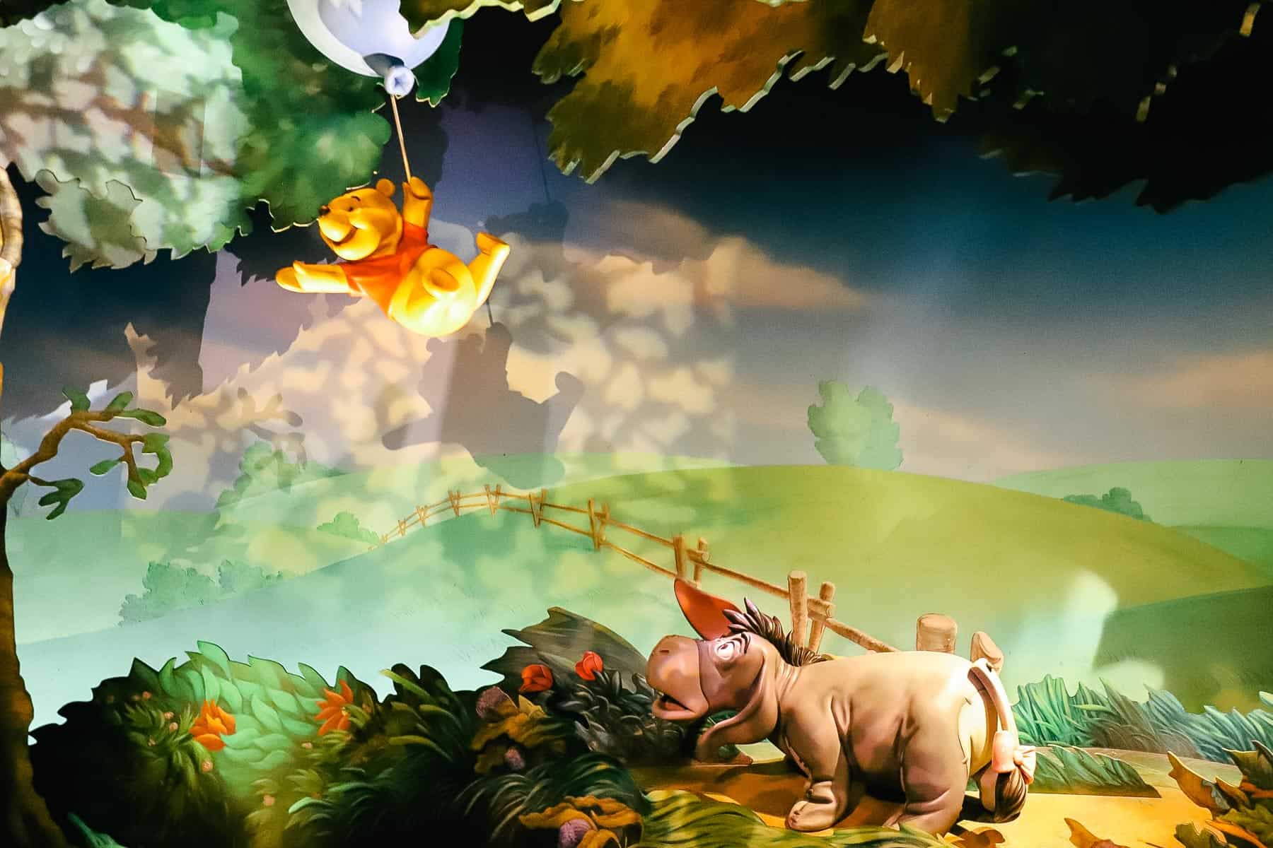 Winnie the Pooh floats to the top of the trees with a balloon while Eeyore looks on from below. 